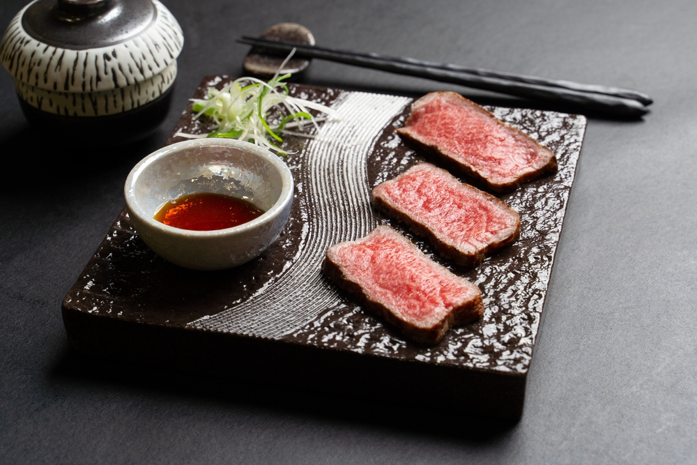 Three slices of wagyu beef on a black slate with chopsticks and soy sauce in a bowl.