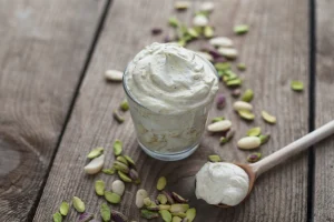Pistachio yoghurt in a glass on wooden slats with pistachio shells and wooden spoon with some yoghurt on it.