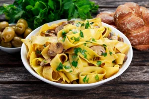 Pappardelle and mushrooms in a white dish on the table with herbs.