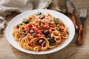 A white dish of pasta puttanesca with cutlery.
